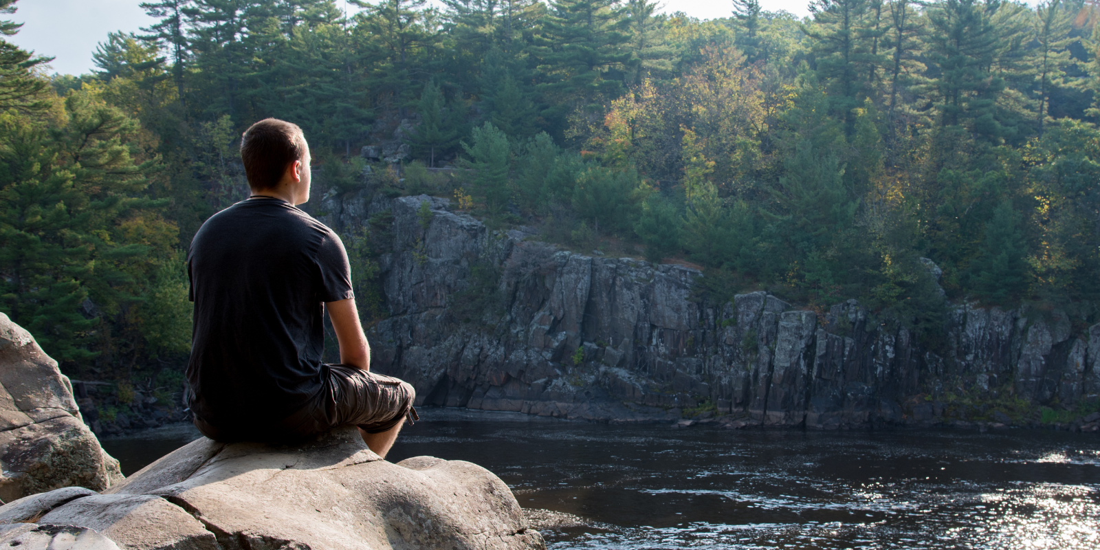 Teenage boy reflecting quietly, sitting on a rock near water.  